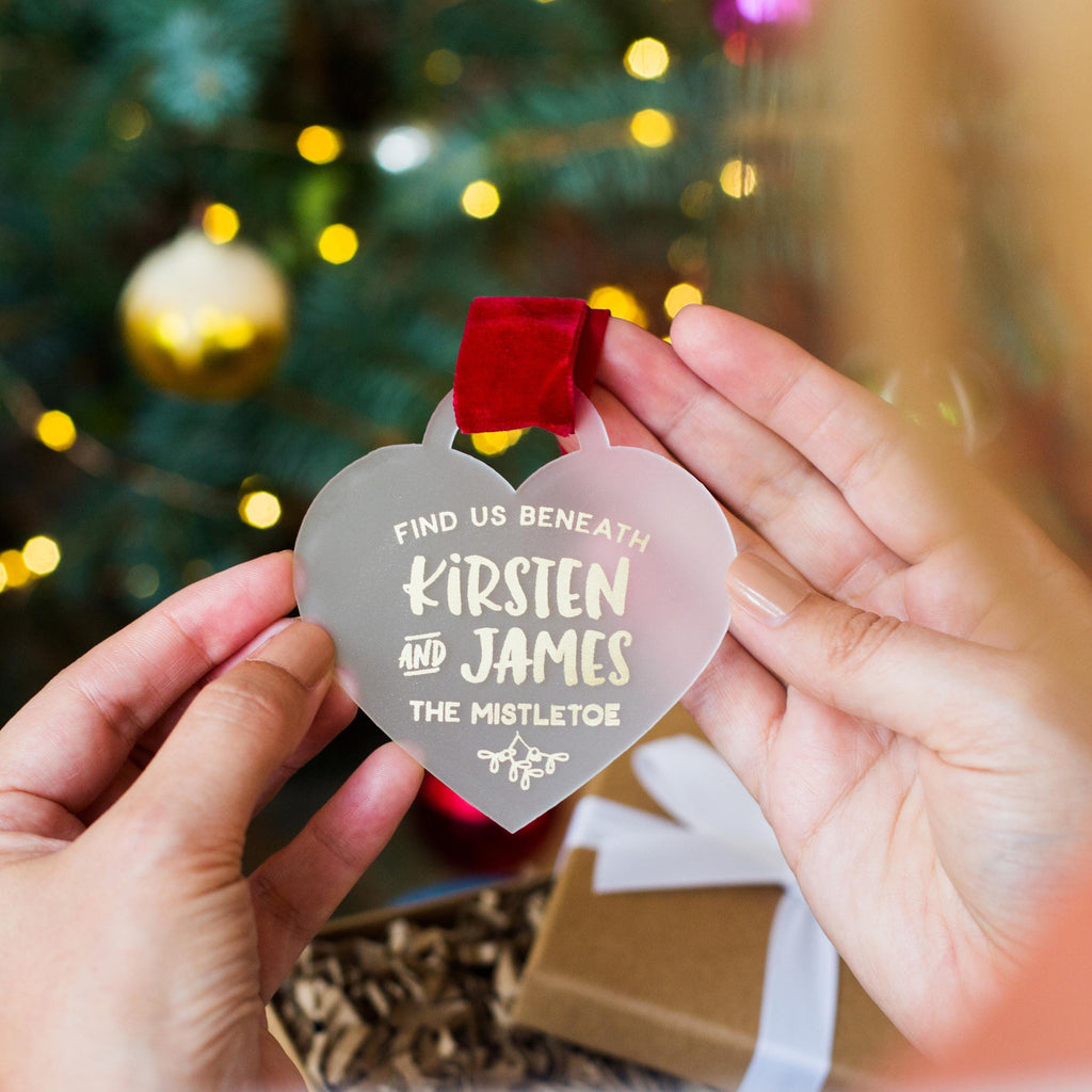 A person's hands holding a heart-shaped ornament with a message on it, in front of a Christmas tree with lights.