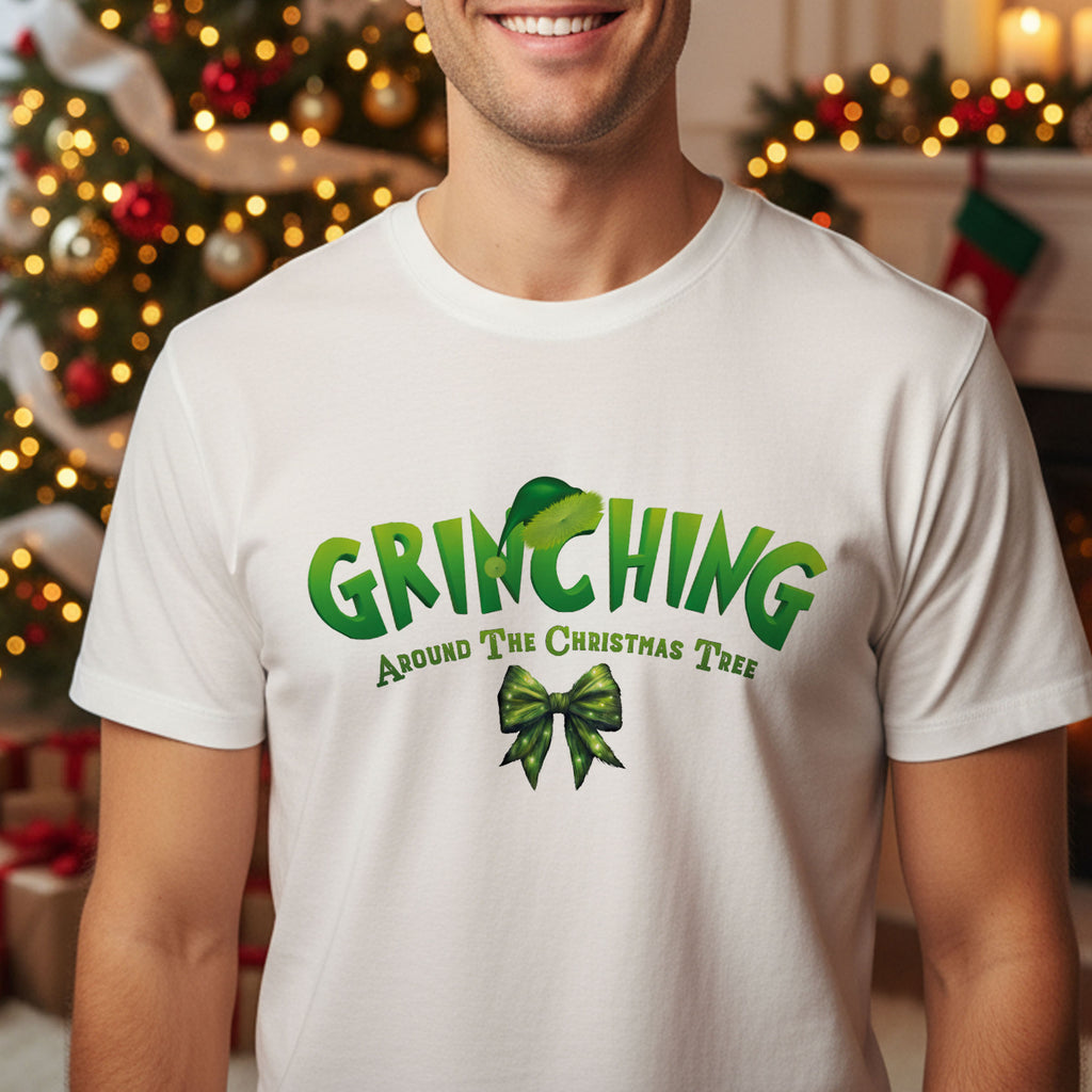 A man wearing a white t-shirt with a graphic design featuring the text "GRINCHING Around The Christmas Tree" and a green bow.