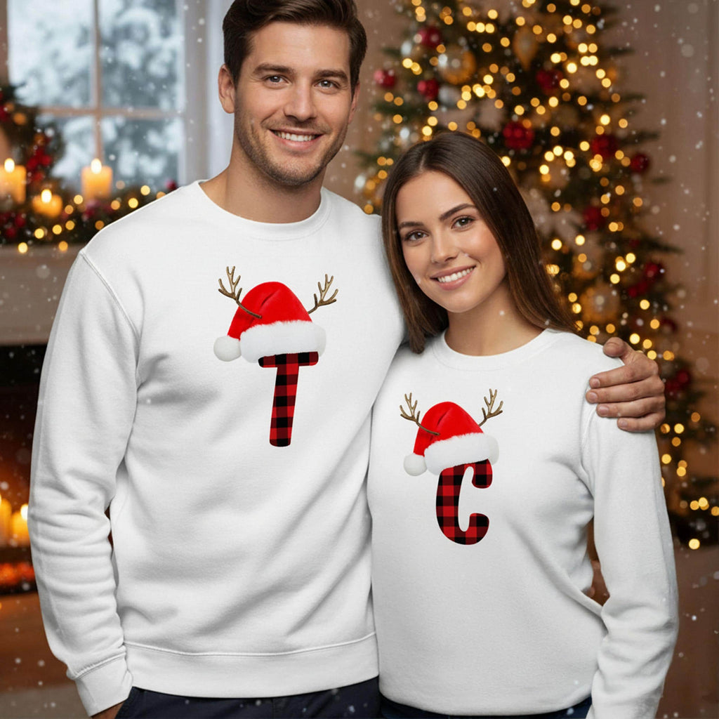 A man and a woman are standing together in front of a Christmas tree, both wearing matching white sweatshirts with a red and black plaid Santa hat and a red letter &#39;C&#39; on them.