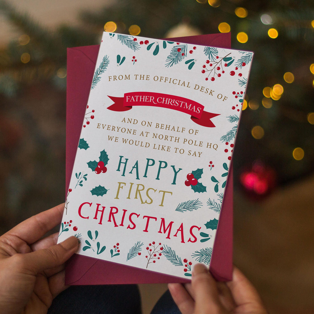 a person holding a Christmas card with a festive design. The card has a message from the official desk of Father Christmas and a message about the first Christmas, with a background of holiday decorations.