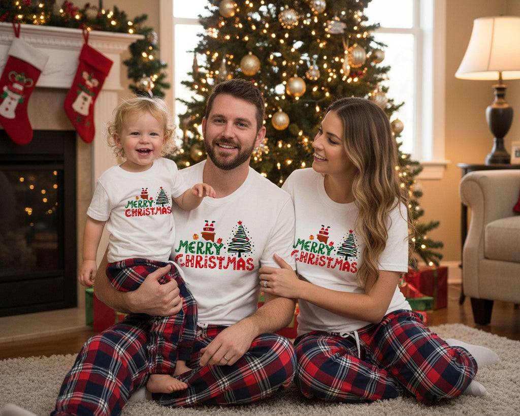 A family of three, consisting of a man, a woman, and a young child, is sitting together in front of a Christmas tree, all wearing matching white t-shirts with the text "Merry Christmas" printed on them.